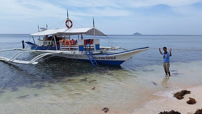 A man pulls a traditional outrigger boat, named "King Indolf," toward a sandy beach, ready for boat tours and island hopping adventures. The vessel is anchored near the shore, with the sea calm under a partly cloudy sky and distant islands visible on the horizon.