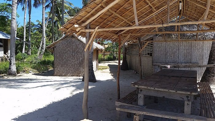 A rustic beachside setting on Araw Beach, with a thatched roof shelter and wooden table under swaying palms. Bamboo huts dot the sandy shore, while lush greenery creates a serene backdrop—perfect for relaxing after a day of island hopping or local boat tours.