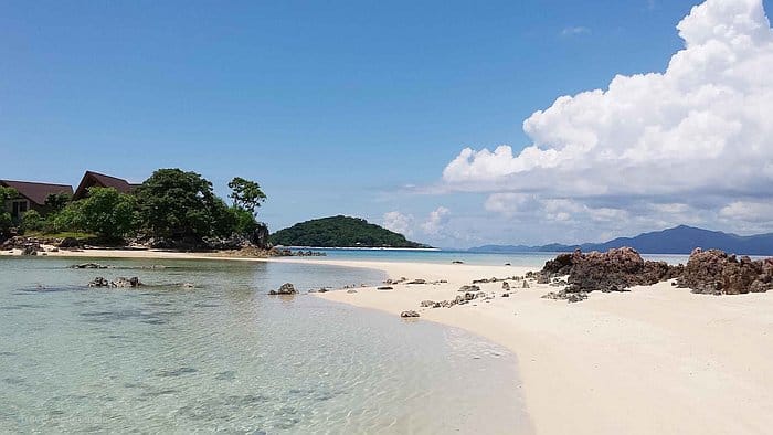 A serene beach with clear turquoise water, white sand, and rocky formations. Lush green hills are visible in the background under a bright blue sky with scattered clouds. A small structure is nestled among the trees on the left, offering a perfect view for island hopping around Bulog Dos.