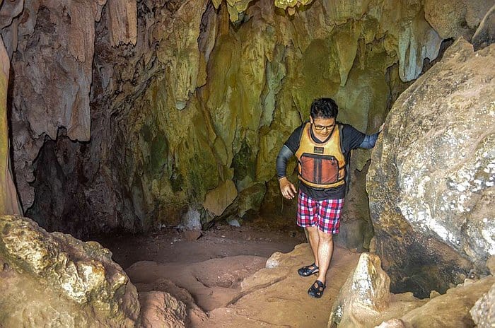 A person wearing a life vest and plaid shorts explores a rocky cave interior near Coron, using one hand for balance against the wall. The cave features rough, textured surfaces and earthy colors, with dim natural lighting reminiscent of Palawan's hidden gems.