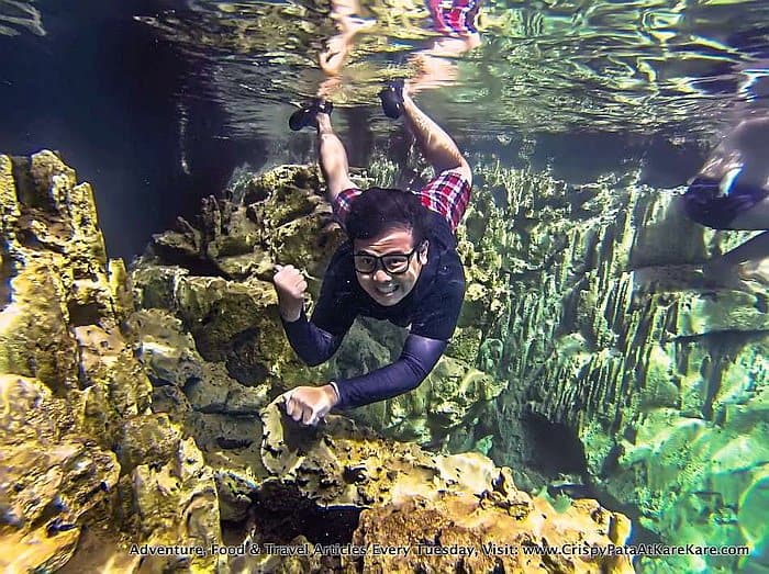 A person wearing goggles and checkered swim trunks is snorkeling underwater near rock formations and coral in the pristine waters of Kayangan Lake, Coron. They seem to be enjoying the exploration of Palawan’s stunning aquatic scenery.