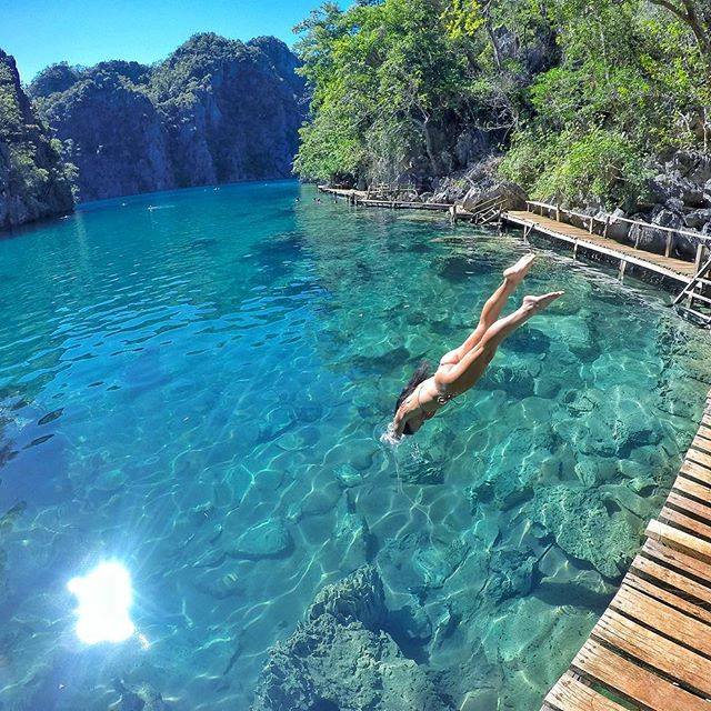 A person dives into the clear blue waters of Kayangan Lake in Palawan, surrounded by lush green trees and rocky cliffs. A wooden dock is visible on the right, with sunlight reflecting off the water's surface. The scene in Coron is bright and serene.