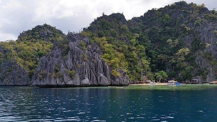 A scenic view of rocky cliffs covered in greenery alongside a calm blue sea evokes the tropical allure of Palawan. Small wooden structures and boats near the tranquil shoreline hint at Coron’s charm, suggesting a remote island village. The partly cloudy sky completes this picturesque scene.