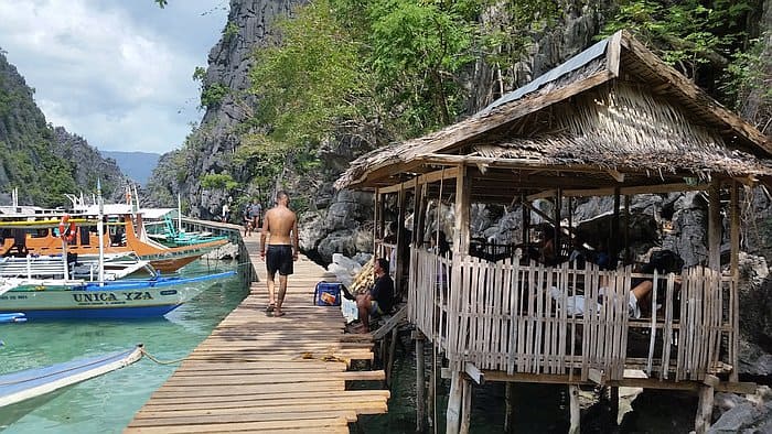 A shirtless man walks on a wooden boardwalk beside the turquoise waters of Palawan, near traditional boats and a bamboo hut. Cliffs and lush greenery surround Kayangan Lake, crafting a stunning tropical landscape.