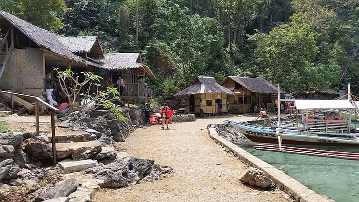 A scenic view of a tropical waterfront in Palawan showcases rustic thatched-roof huts surrounded by lush greenery. Nearby, a small group gathers as a traditional boat rests in the clear turquoise water, reminiscent of Coron near the famed Kayangan Lake.