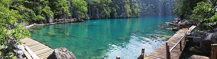 A serene scene at Kayangan Lake in Coron, Palawan, where lush greenery and rocky cliffs embrace the water. Wooden pathways lead to the edge, as a person in a canoe paddles across the clear, turquoise water under a bright sky.