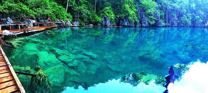 A stunning lagoon reminiscent of Kayangan Lake in Coron, with crystal-clear turquoise water, surrounded by lush green foliage and rock formations. A wooden pathway extends over the water, blending harmoniously with the natural beauty of Palawan.