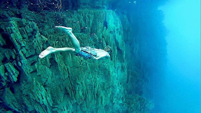 A person wearing swim shorts is free diving underwater near a rocky cliff in Coron, surrounded by the clear blue waters of Kayangan Lake. The rocks have sharp, jagged edges, and the scene conveys a sense of adventure and exploration in the heart of Palawan.