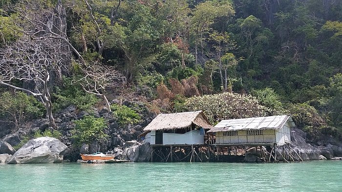 A small, rustic hut with a thatched roof stands on stilts over turquoise water, nestled against a lush, rocky hillside. Perfect for island hopping adventures, a wooden boat is moored nearby, with dense greenery and some bare trees in the background.