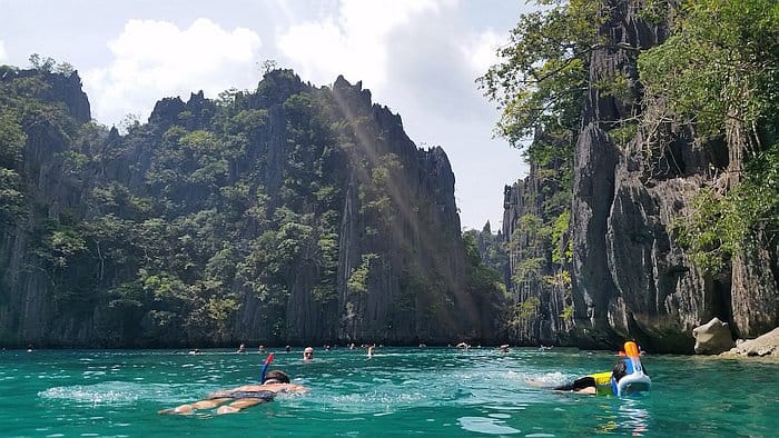 People snorkeling in the clear turquoise waters of Coron are enveloped by rocky cliffs and lush greenery under a partly cloudy sky, turning this picturesque excursion into an unforgettable memory.