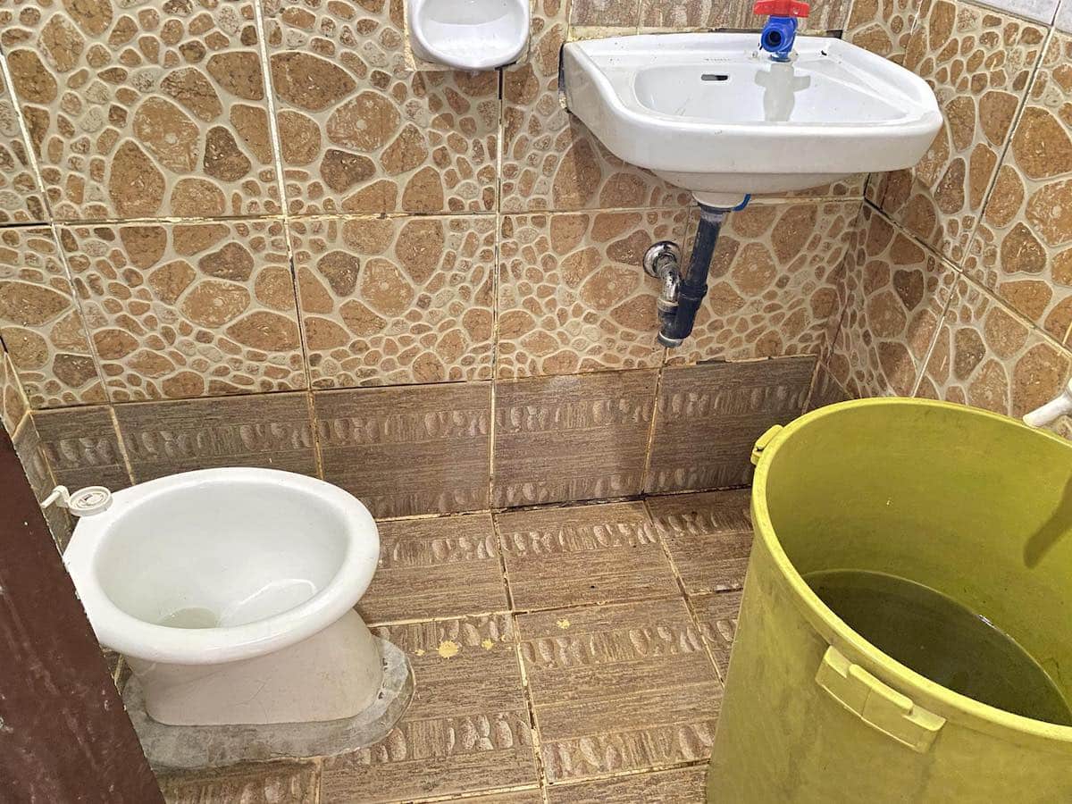 A small bathroom reminiscent of a cozy Banana Island retreat features a white squat toilet, a wall-mounted sink, and a large yellow bucket on tiled floors. The walls are adorned with brown patterned tiles, evoking the rustic charm of an island-hopping adventure.