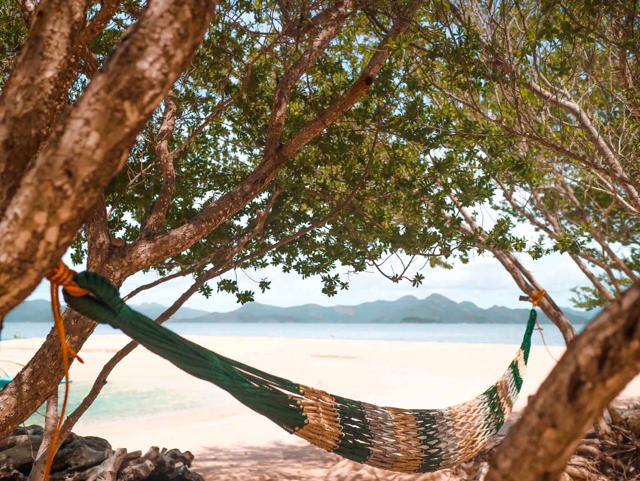 A colorful hammock is suspended between trees on a tropical beach, perfect for island hopping adventures. The shoreline stretches into clear blue waters, with mountains visible in the hazy distance under a sunny sky. Nearby, Banana Island awaits explorers eager for boat tours.