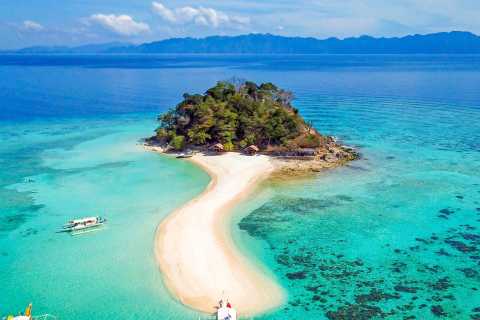 A small, lush haven, Banana Island is surrounded by clear turquoise water and connected by a sandy beach path. Nearby boats hint at island hopping adventures, while distant mountains rise under a clear blue sky.