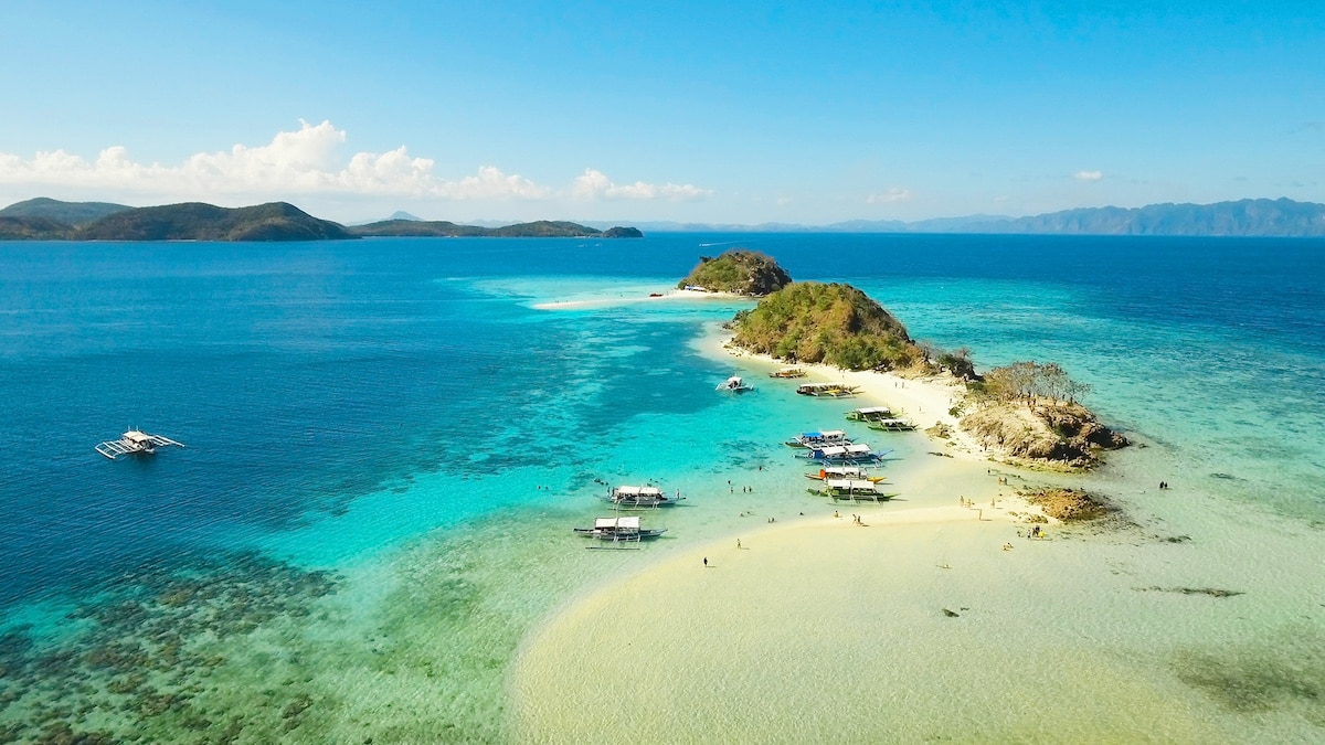 A picturesque aerial view of Banana Island showcases sandy beaches surrounded by clear, turquoise waters. Several small boats, ready for island hopping and boat tours, are docked near the shore, while lush greenery adorns the hilly landscape. Distant islands linger on the horizon.