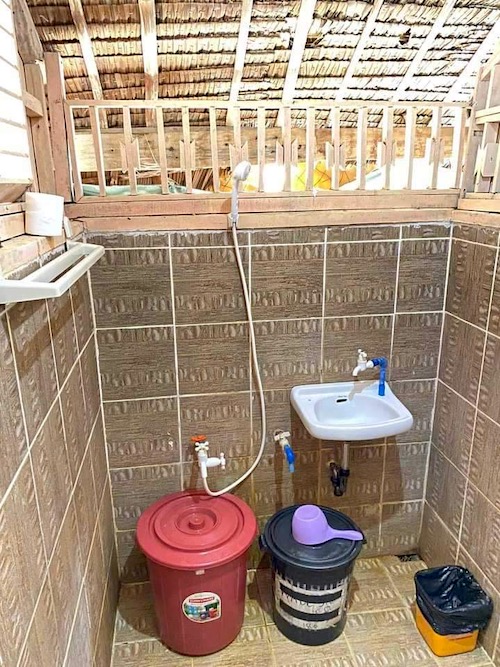 A rustic bathroom reminiscent of an island escape, featuring brown patterned tiles, a small white sink, and an overhead shower. Two buckets—one red and one black with a ladle—sit next to a small waste bin on the tiled floor. The ceiling evokes Banana Island with its thatched roofing.