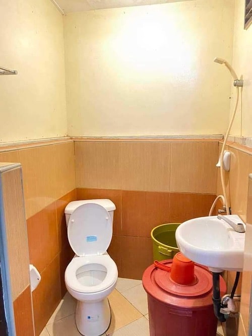 A small bathroom with beige and tan tile walls, ideal for a refreshing rinse after island hopping. It features a toilet, white sink, showerhead, and two buckets—one green and one red with a lid. The floor is adorned with light-colored tiles.