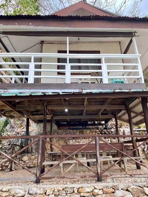 A two-story structure with a white railing on the upper balcony, set against a rustic stone foundation on the lower level, makes for an ideal retreat after island hopping. The bottom porch features wooden beams and a stone wall, surrounded by rocks and sparse foliage.