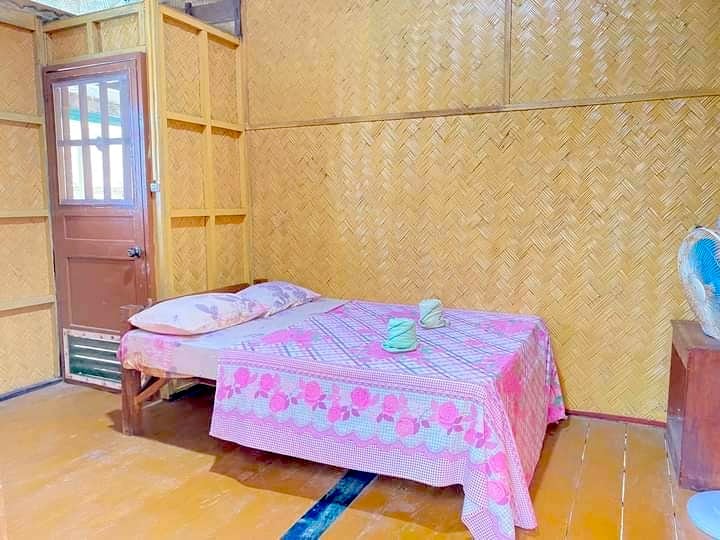 A simple room with wooden woven walls features a bed covered in a pink floral blanket and two pillows. A wooden door with a glass window is in the background, hinting at adventures like island hopping or boat tours around Banana Island, while a small fan rests on a table beside the bed.