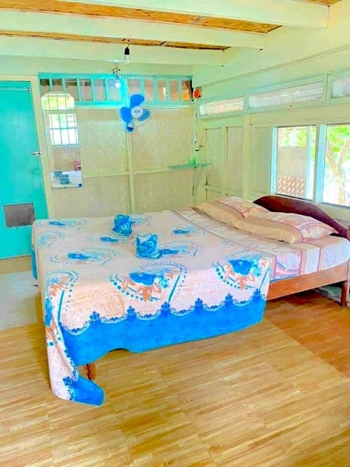 A cozy bedroom with a wooden floor, featuring a double bed adorned with a colorful floral bedspread. A blue fan is mounted on the wall, evoking the refreshing breezes of island hopping adventures. The room has light turquoise accents, and windows that let in natural light like Banana Island's shores.