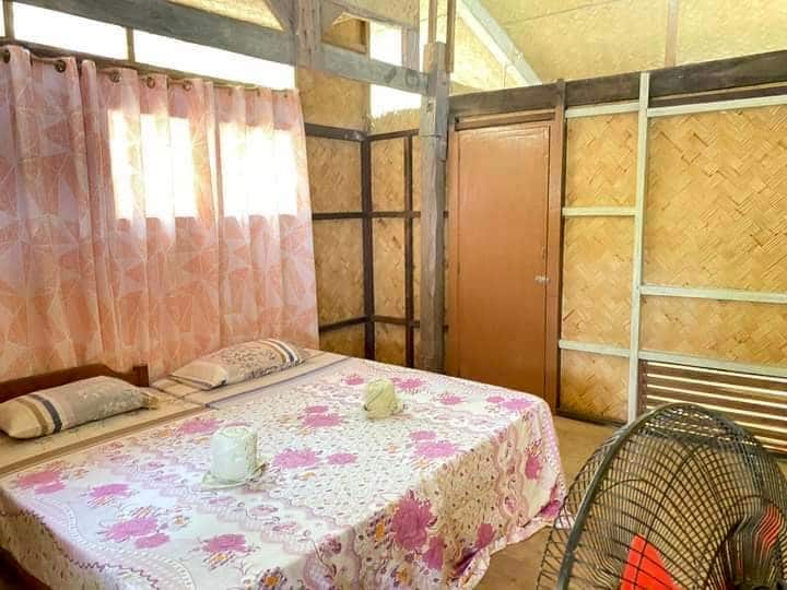 A cozy room with wooden walls and a thatched section evokes the charm of Banana Island. It features a bed with floral bedding, two pillows, and a fan at the foot. A pink patterned curtain covers the window, inviting guests to relax between island hopping adventures.