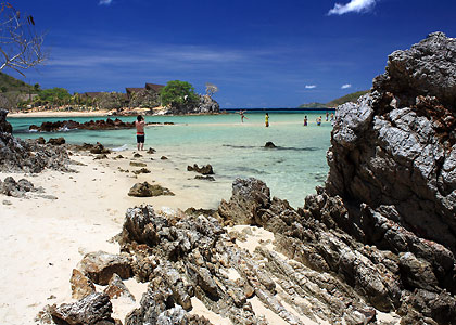 A tropical beach scene at Bulog Dos features clear turquoise water and rocky formations. People are enjoying island hopping, swimming, and standing in the shallow waters. The sky is bright blue with a few clouds, and a thatched-roof building completes the tranquil backdrop.