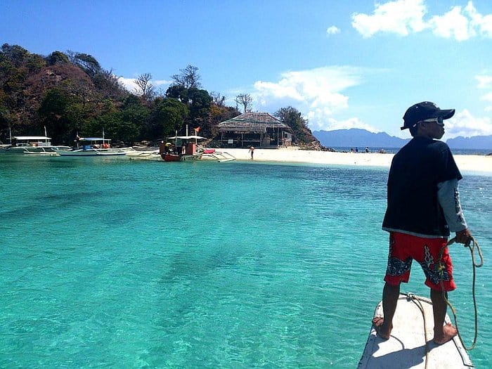 A person stands on a boat wearing a hat and sunglasses, overlooking the tropical island of Bulog Dos with clear turquoise water. The island has a sandy beach, trees, and a few buildings. Nearby, boats are anchored under the sunny blue sky—a perfect spot for island hopping and boat tours.