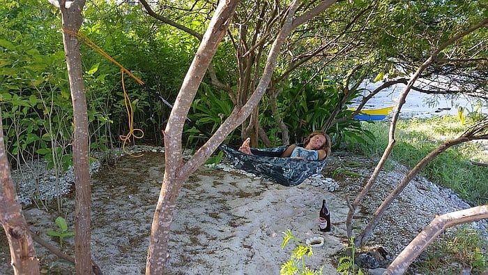 A person relaxes in a hammock tied between trees, surrounded by lush greenery. There is a bottle on the ground beside them. Sunlight filters through the leaves, creating a peaceful, shaded spot.