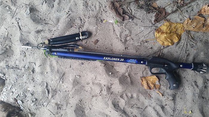 A speargun labeled "Explorer 20" lies on sandy ground. It has a blue body and a black handle. Nearby are some dry leaves and an object resembling a knife or tool sheath in black.