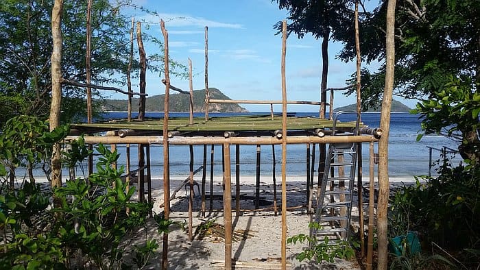 A partially constructed bamboo structure stands on a sandy beach, surrounded by trees. A ladder leans against the frame. In the background, there is a view of the ocean and a distant island under a clear blue sky.