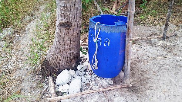 A blue plastic barrel used for collecting rainwater is tied to a tree trunk with a rope. It is situated on sandy ground, surrounded by greenery and a few white stones.