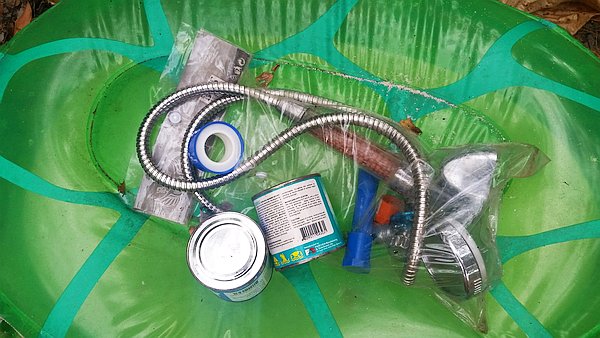A green inflatable pool holds various plumbing tools and supplies, including a coiled metal hose, adhesive tape, a metal showerhead, plastic tubing, and two small containers. Items are in clear plastic packaging.