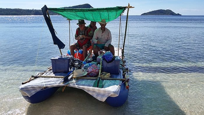 Three people sit on a small, makeshift boat with a green canopy, floating in shallow, clear water. The boat is loaded with supplies. In the background, a scenic view of the ocean and distant islands can be seen.