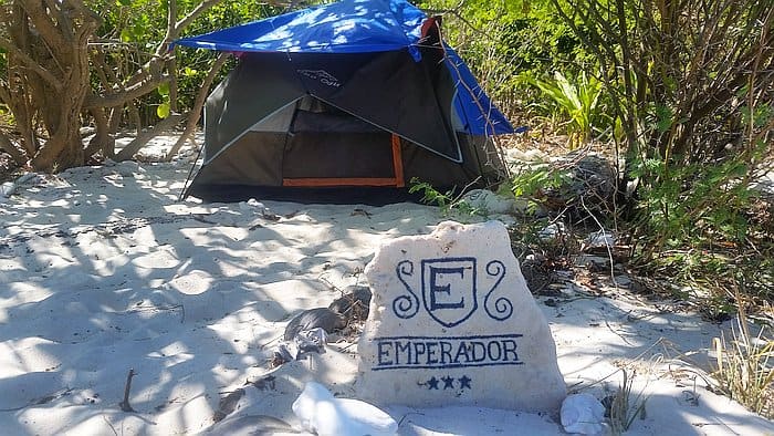 A blue and gray tent is set up on a sandy beach surrounded by greenery. In the foreground, there's a stone with an emblem and the word "EMPERADOR" engraved on it.