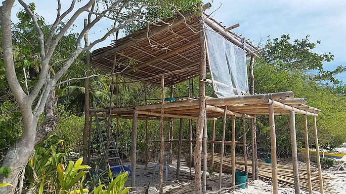 A wooden structure on a sandy beach surrounded by lush greenery. It consists of bamboo poles and thatched roofs, partially covered by a white tarp. A ladder and construction materials are visible nearby.