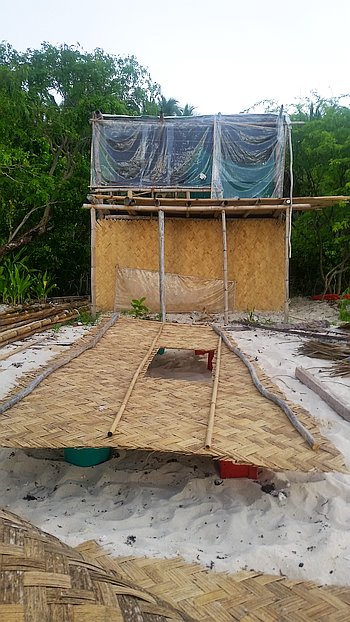 A small makeshift bamboo and tarp shelter on a sandy beach, surrounded by green trees. A narrow woven bamboo path leads up to the structure, and colorful containers are visible underneath.