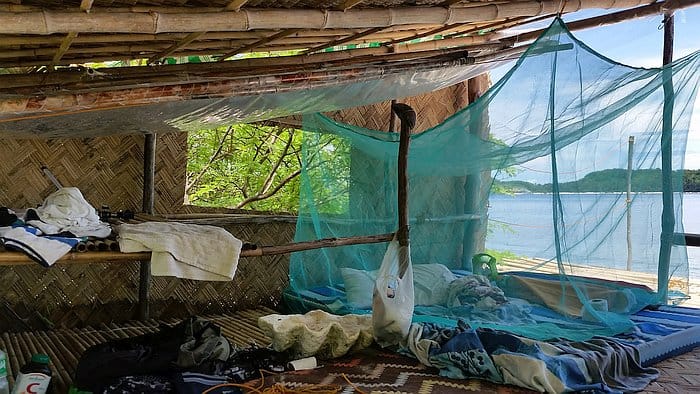 A rustic, open-air bamboo hut with a thatched roof overlooking a serene body of water. Inside, there's a makeshift bed with a blue mosquito net, clothes, and personal items scattered around. The view includes lush greenery and clear skies.