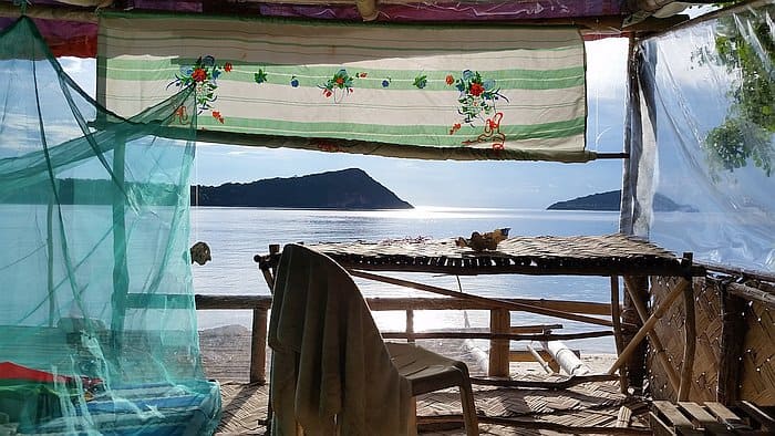 A rustic bamboo hut overlooking a serene beach with calm blue water. Inside, a simple chair and table are under a colorful fabric roof, with a green mosquito net hanging nearby. Two distant islands are visible under a bright sky.