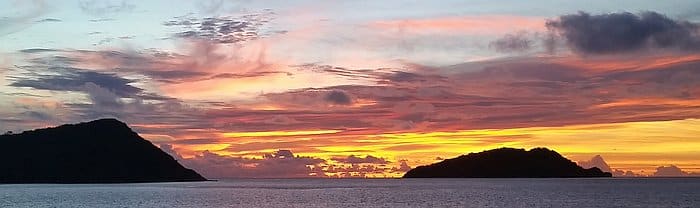 A serene sunset over the ocean with vibrant orange and pink hues in the sky. Silhouettes of two islands are visible on the horizon, creating a peaceful and picturesque scene.