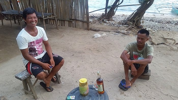 Two men are sitting outdoors on wooden stools by the beach, enjoying a sunny day after some island hopping. One is smiling in a white shirt, while the other wears green. Between them, a small table holds a bottle, a jar, and some items. A rustic wooden fence lines the background.