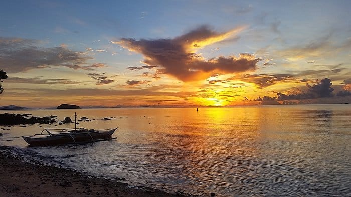 A small boat, perhaps poised for island hopping, rests on a rocky shore at sunset. The sky is painted with vibrant orange and yellow hues, reflecting on the calm sea. Fluffy clouds drift across the horizon, and an island is visible in the distance.