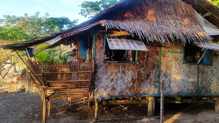 A rustic bamboo hut with a thatched roof stands in a sunlit area, perfect for island hopping adventures. It features a small raised porch with wooden railings. Trees and greenery are visible in the background as the hut is elevated slightly above the ground, offering a serene escape.