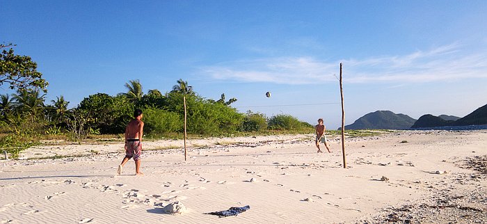 Two people playing beach volleyball on a sandy shore under a clear blue sky, surrounded by lush greenery and distant hills, while the gentle rhythm of waves hints at an inviting boat trip just beyond the horizon.