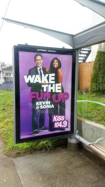Bus stop advertisement featuring two smiling individuals, a man in a suit and glasses and a woman in a black dress. They stand under bold text reading "Wake the Fun Up," highlighting their tour excitement with "Kevin & Sonia" and "Kiss 104.9" displayed at the bottom.