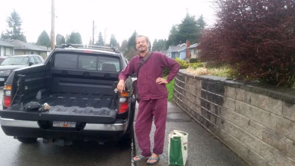 A person in a purple outfit stands smiling next to a pickup truck on a rainy day, perhaps ready for a road trip. They have one hand on the truck and the other on their hip. A reusable shopping bag is on the ground beside them. A brick wall and houses line the street.