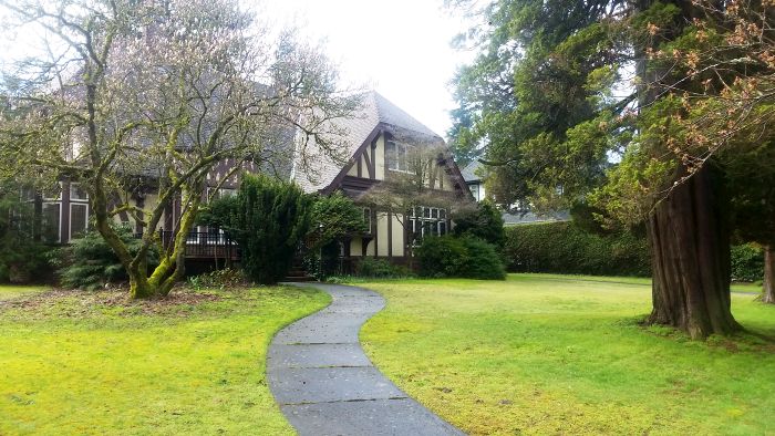 A quaint Tudor-style house stands in a lush green yard, perfect for a scenic tour. It features wooden beams and a steeply pitched roof. A curved stone path leads to the entrance, flanked by tall trees and shrubs. The overcast sky enhances the serene atmosphere of this picturesque spot.