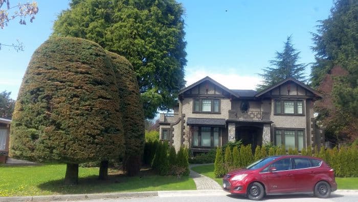 A two-story house with a brown exterior and large windows is surrounded by greenery, inviting visitors on a picturesque tour. Neatly trimmed bushes and a large tree adorn the yard, while a red car, ready for the next road trip adventure, rests on the street in front of the house.