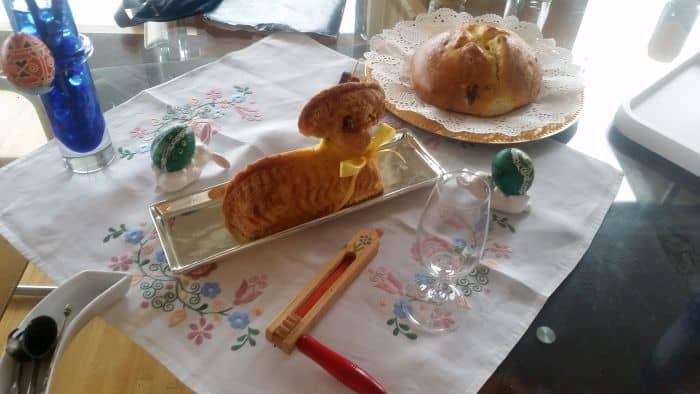 A festive table reminiscent of a delightful tour, featuring a lamb-shaped cake, a round loaf of bread, painted eggs on stands, and a blue glass vase. Adorned with a floral embroidered cloth, it also holds an inviting glass and wooden-handled tool.