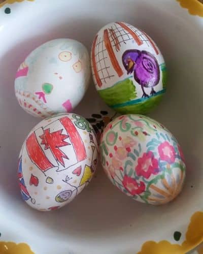 Four intricately decorated eggs rest in a white bowl with a yellow rim, as if they’ve been collected on a vibrant road trip. Each egg features unique, colorful designs, including geometric patterns, floral motifs, and a bird illustration.