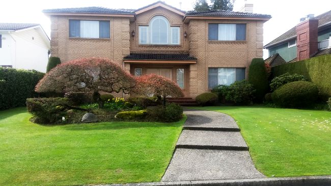 A two-story brick house with a manicured lawn and a curved walkway leading to the entrance awaits your tour. The front yard features well-trimmed bushes and trees, making it a perfect stop on your road trip.