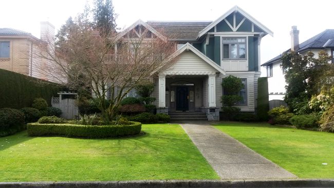 A two-story house with a gabled roof and covered porch invites visitors from the pathway, flanked by trees and shrubs, perfect for a road trip tour. Neatly maintained lawn and trimmed hedges surround it, adding to its charming allure.